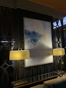 A modern interior space featuring a large abstract painting with shades of blue and white, mounted on a dark patterned wall. The painting is flanked by two elegant lamps with beige lampshades, emitting a warm light that softly illuminates the area. The background consists of decorative wall panels with an interlocking circular design.