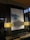 A modern interior space featuring a large abstract painting with shades of blue and white, mounted on a dark patterned wall. The painting is flanked by two elegant lamps with beige lampshades, emitting a warm light that softly illuminates the area. The background consists of decorative wall panels with an interlocking circular design.