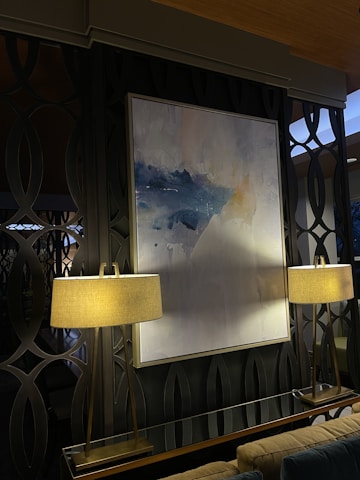 A modern interior space featuring a large abstract painting with shades of blue and white, mounted on a dark patterned wall. The painting is flanked by two elegant lamps with beige lampshades, emitting a warm light that softly illuminates the area. The background consists of decorative wall panels with an interlocking circular design.