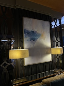A modern interior space featuring a large abstract painting with shades of blue and white, mounted on a dark patterned wall. The painting is flanked by two elegant lamps with beige lampshades, emitting a warm light that softly illuminates the area. The background consists of decorative wall panels with an interlocking circular design.