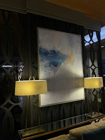 A modern interior space featuring a large abstract painting with shades of blue and white, mounted on a dark patterned wall. The painting is flanked by two elegant lamps with beige lampshades, emitting a warm light that softly illuminates the area. The background consists of decorative wall panels with an interlocking circular design.