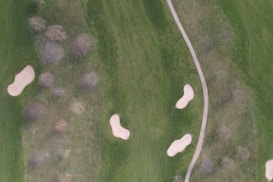An aerial view of a golf course featuring a green landscape with multiple sand traps and a paved pathway. Trees with bare branches are scattered throughout the area, indicating the season might be winter or early spring. The scene is serene and well-maintained, typical of a professional golf course.