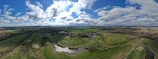 A sprawling landscape with a lush green golf course featuring several sand bunkers and water hazards, surrounded by open fields and trees. In the distance, a small village or town with clusters of houses is visible. The sky is filled with fluffy clouds scattered across a bright blue backdrop, creating a serene and expansive view.
