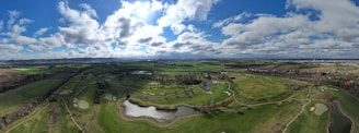 A sprawling landscape with a lush green golf course featuring several sand bunkers and water hazards, surrounded by open fields and trees. In the distance, a small village or town with clusters of houses is visible. The sky is filled with fluffy clouds scattered across a bright blue backdrop, creating a serene and expansive view.