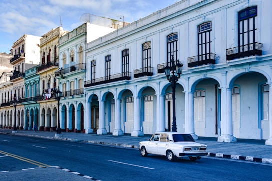 A street lined with historic buildings featuring large archways and intricate balconies. The pastel-colored facades exhibit an array of architectural details, reflecting a colonial style. A vintage white car is parked on the road, enhancing the scene's nostalgic atmosphere. The street is empty, creating a tranquil and timeless impression.