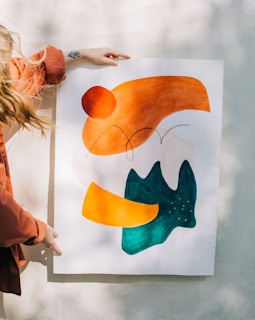 A person holds a piece of abstract artwork featuring bold shapes and colors, including orange and teal elements on a white background. The figure in the image is partially visible, showing a tattoo on their wrist.