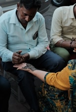 A person attentively applies intricate henna designs on another person's hand. The scene captures the focus and skill involved in creating detailed body art. Both individuals are seated, with various elements of casual clothing visible.