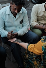 A person attentively applies intricate henna designs on another person's hand. The scene captures the focus and skill involved in creating detailed body art. Both individuals are seated, with various elements of casual clothing visible.