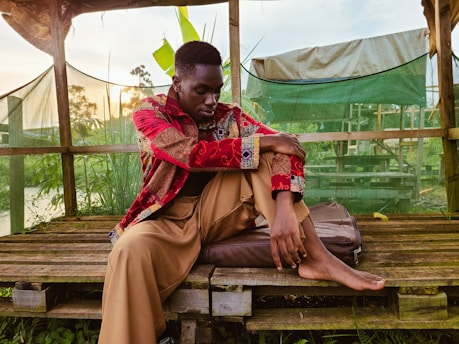 A person sits on a wooden platform surrounded by green foliage. They are wearing a vibrant red and patterned shirt with beige pants, sitting with one knee up and appearing deep in thought. Behind them, there is a canopy and some netting used for shade, with a glimpse of the natural landscape in the background.