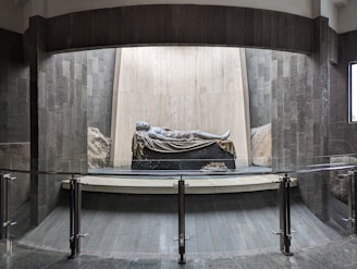A stone sculpture of a reclining figure is set on a black base, surrounded by a modern architectural setting with textured dark stone walls and a glass railing in the foreground. The background features a mix of rough, natural stone and smoother, light-colored surfaces that create a contrast with the darker elements.
