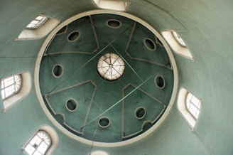 A circular ceiling with a central decorative element, surrounded by rectangular windows. The ceiling is painted in a soft green color with symmetrical architectural details and round openings. The lighting is soft, highlighting the textures and curves of the structure.