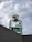 A glass perfume bottle labeled 'Louis Cardin Adventure' is prominently positioned on a ledge, with a cloudy sky in the background. The bottle cap has a metallic finish and the liquid inside is a light turquoise color.