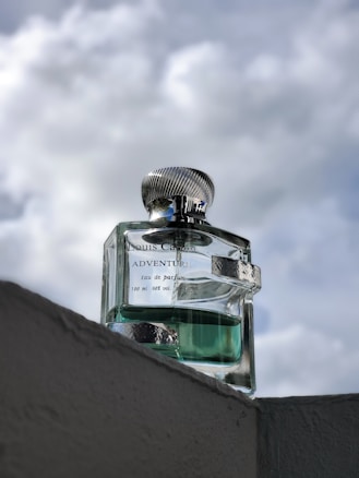 A glass perfume bottle labeled 'Louis Cardin Adventure' is prominently positioned on a ledge, with a cloudy sky in the background. The bottle cap has a metallic finish and the liquid inside is a light turquoise color.