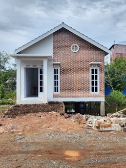A small house with a combination of red brick facade and white walls, elevated on stilts. The house features a gable roof and multiple windows, with a small entrance at the front. The construction site in the foreground has piles of bricks and dirt.