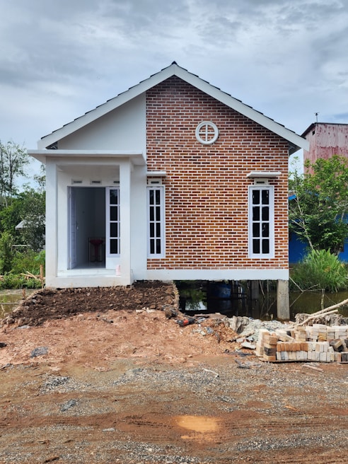 A small house with a combination of red brick facade and white walls, elevated on stilts. The house features a gable roof and multiple windows, with a small entrance at the front. The construction site in the foreground has piles of bricks and dirt.