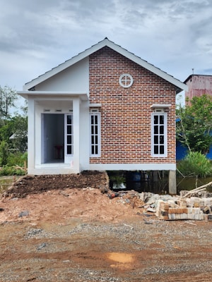A small house with a combination of red brick facade and white walls, elevated on stilts. The house features a gable roof and multiple windows, with a small entrance at the front. The construction site in the foreground has piles of bricks and dirt.