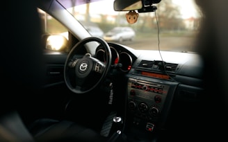 Interior view of a car focusing on the steering wheel and dashboard. The Mazda logo is visible on the steering wheel. The dashboard features various controls and a digital display, with a hanging air freshener in the shape of a cute monkey. The image conveys a sense of warmth, with sunlight streaming through the windows.