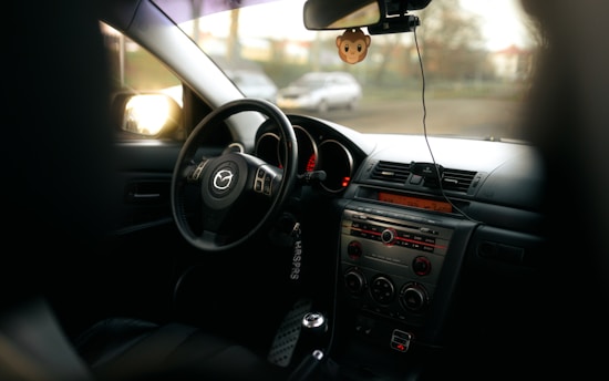 Interior view of a car focusing on the steering wheel and dashboard. The Mazda logo is visible on the steering wheel. The dashboard features various controls and a digital display, with a hanging air freshener in the shape of a cute monkey. The image conveys a sense of warmth, with sunlight streaming through the windows.