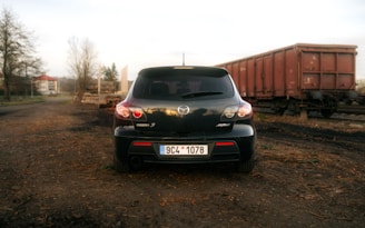 A black Mazda 3 hatchback is parked on a dirt path, with an empty railway track and a couple of rusted train carriages in the background. The scene is set in a somewhat rural area with sparse trees and a distant building visible under an overcast sky.