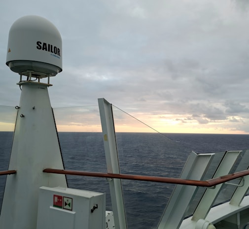 A view of the ocean seen from a ship's deck, featuring a piece of maritime equipment with the word 'Sailor' on it. The horizon is visible with a cloudy sky illuminated by the setting or rising sun, casting a warm glow over the sea. Glass panels and wooden railings are part of the ship's structure.