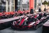 A row of red and black go-karts is parked on a racing track. The karts have numbers and branding on them and are lined up ready for use. In the background, there are more go-karts and a building with glass walls.