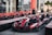 A row of red and black go-karts is parked on a racing track. The karts have numbers and branding on them and are lined up ready for use. In the background, there are more go-karts and a building with glass walls.