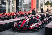 A row of red and black go-karts is parked on a racing track. The karts have numbers and branding on them and are lined up ready for use. In the background, there are more go-karts and a building with glass walls.