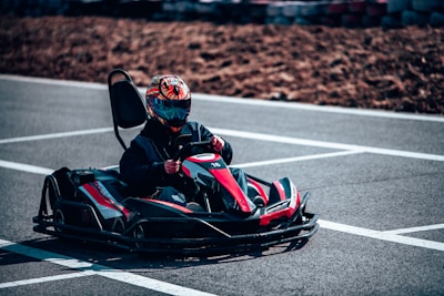 A person is driving a go-kart on an outdoor track. The driver is wearing a colorful helmet and dark clothing. The go-kart is black with red accents and is captured in motion on the gray asphalt track. Surrounding the track are some blurred tire barriers and a brown dirt area.