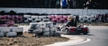 A person is driving a go-kart on a track lined with stacked used tires. The go-kart is turning a corner on the paved road, and the driver is wearing a helmet with colorful designs. The tires used as barriers are painted in various colors, including white, black, and pink.