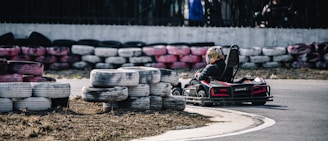 A person is driving a go-kart on a track lined with stacked used tires. The go-kart is turning a corner on the paved road, and the driver is wearing a helmet with colorful designs. The tires used as barriers are painted in various colors, including white, black, and pink.