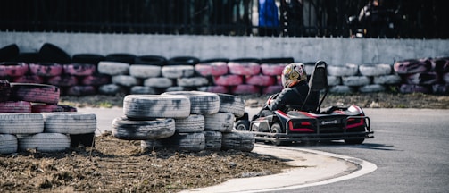 A person is driving a go-kart on a track lined with stacked used tires. The go-kart is turning a corner on the paved road, and the driver is wearing a helmet with colorful designs. The tires used as barriers are painted in various colors, including white, black, and pink.