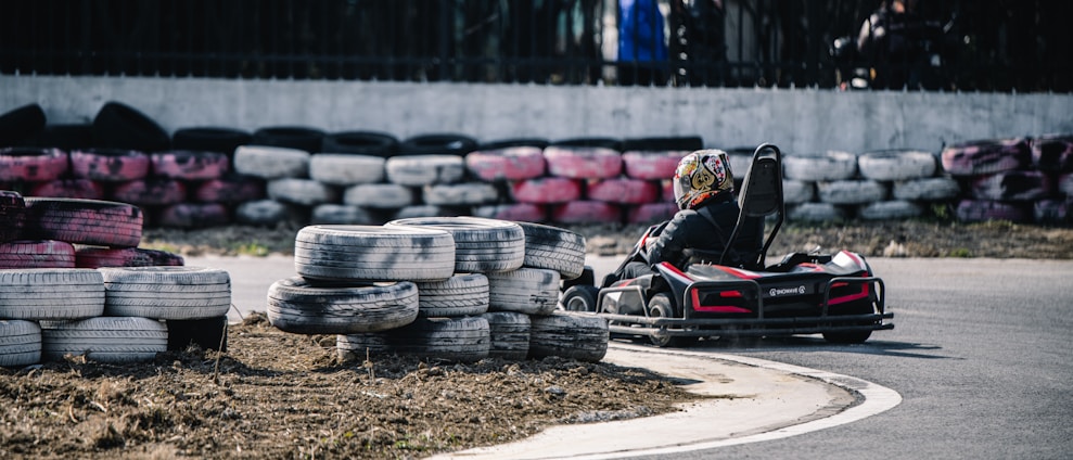 A person is driving a go-kart on a track lined with stacked used tires. The go-kart is turning a corner on the paved road, and the driver is wearing a helmet with colorful designs. The tires used as barriers are painted in various colors, including white, black, and pink.