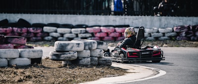 A person is driving a go-kart on a track lined with stacked used tires. The go-kart is turning a corner on the paved road, and the driver is wearing a helmet with colorful designs. The tires used as barriers are painted in various colors, including white, black, and pink.
