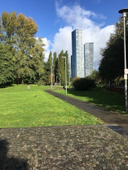 A pathway winds through a lush green park with trees on either side. In the background, two tall modern skyscrapers rise against a partly cloudy blue sky. The foreground features a stone-tiled area, while a bird can be seen on the grass.