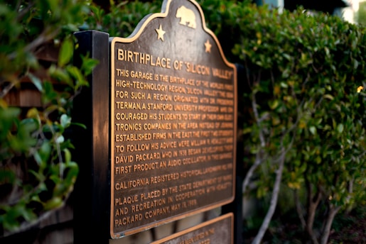 A plaque stands amidst greenery, featuring an inscription about the birthplace of Silicon Valley. The text highlights the historical significance of a garage in the development of the high-technology region.