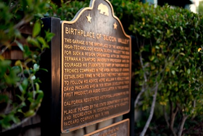 A plaque stands amidst greenery, featuring an inscription about the birthplace of Silicon Valley. The text highlights the historical significance of a garage in the development of the high-technology region.