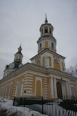 A yellow and white Orthodox church with two onion domes featuring crosses at the top. The building is surrounded by snow and there is a green fence partially visible in front. The sky is overcast, giving the scene a grey backdrop.