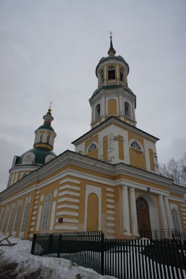 A yellow and white Orthodox church with two onion domes featuring crosses at the top. The building is surrounded by snow and there is a green fence partially visible in front. The sky is overcast, giving the scene a grey backdrop.