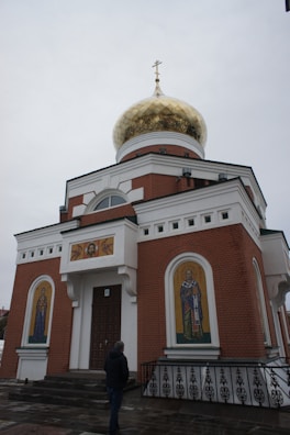 A small brick orthodox church features a large golden onion dome topped with a cross. The facade includes religious icons and paintings, with a single person standing in front. The overcast sky creates a somber and reflective atmosphere.