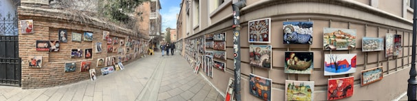 A street lined with brick and stucco walls displays numerous colorful paintings. The artwork is hung on both sides of the narrow path, creating a vibrant outdoor gallery. People are walking and observing the art, adding life to the scene. The setting feels urban, possibly in a historic district.