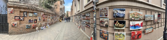 A street lined with brick and stucco walls displays numerous colorful paintings. The artwork is hung on both sides of the narrow path, creating a vibrant outdoor gallery. People are walking and observing the art, adding life to the scene. The setting feels urban, possibly in a historic district.