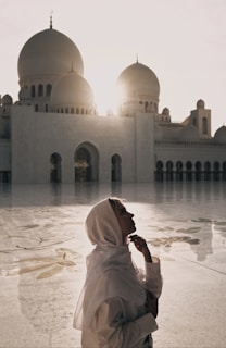 A person wearing a light-colored headscarf stands in front of a grand mosque with large domes. The sun is setting or rising behind the mosque, casting a warm glow and long shadows on the intricately patterned courtyard.