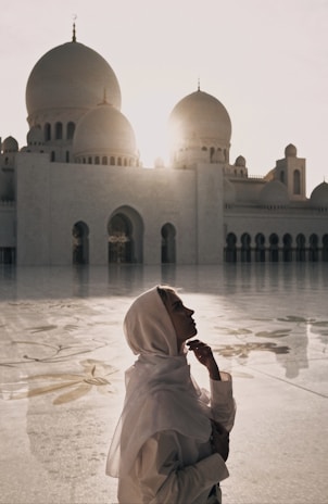 A person wearing a light-colored headscarf stands in front of a grand mosque with large domes. The sun is setting or rising behind the mosque, casting a warm glow and long shadows on the intricately patterned courtyard.