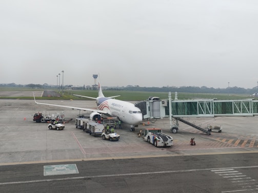 A commercial airplane is parked at the airport gate, connected to a jet bridge. Ground service vehicles, including baggage carts and fuel trucks, are positioned around the aircraft. The runway and a control tower are visible in the background, along with a cloudy sky.