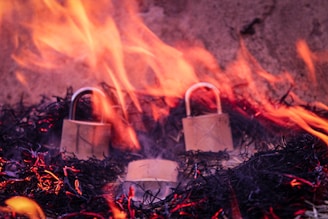 Two metallic padlocks are surrounded by flames and embers on a dark background. The fire creates an intense and dramatic effect as it engulfs the locks.