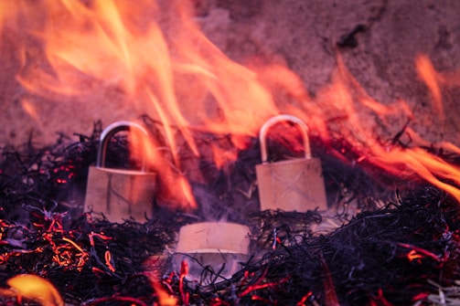 Two metallic padlocks are surrounded by flames and embers on a dark background. The fire creates an intense and dramatic effect as it engulfs the locks.