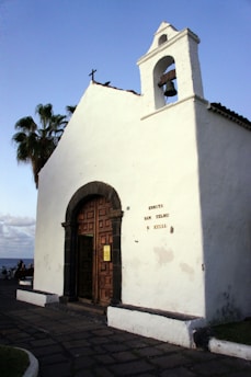 A white, small, historic chapel with a wooden door and an attached bell tower. The building has some inscriptions on its facade and is partially covered by shadows. In the background, there are palm trees and a clear blue sky.