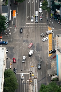 An aerial view of a busy urban intersection with multiple cars in motion, surrounded by tall buildings and visible trees at the edge. Pedestrians can be seen at the crosswalks and several vehicles, including a red car and a white van, navigate through the various lanes. Traffic lights and road markings guide the flow of vehicles.
