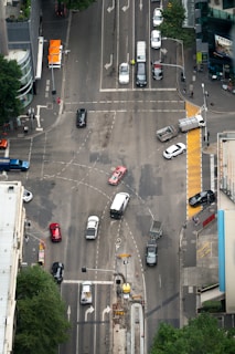 An aerial view of a busy urban intersection with multiple cars in motion, surrounded by tall buildings and visible trees at the edge. Pedestrians can be seen at the crosswalks and several vehicles, including a red car and a white van, navigate through the various lanes. Traffic lights and road markings guide the flow of vehicles.