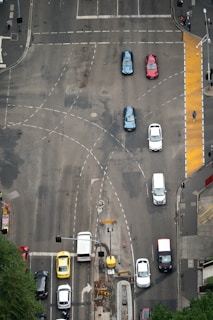 An aerial view of a busy urban intersection with multiple cars stopped at traffic lights. The intersection is marked with various lanes and dotted lines, and there is a distinct yellow pedestrian path on one side. Several vehicles of different colors, including blue, red, and white, are visible. Pedestrians can be seen on the sidewalks, and there are traffic signals and streetlights visible.